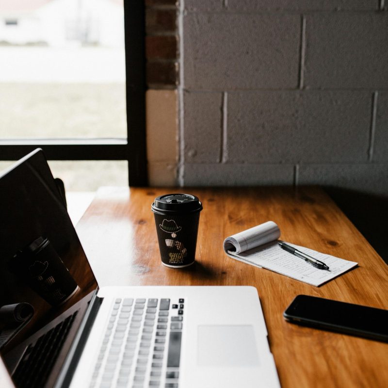 pexels-photo-1967535-1967535 Modern office desk setup with laptop, coffee cup, and notepad by window.